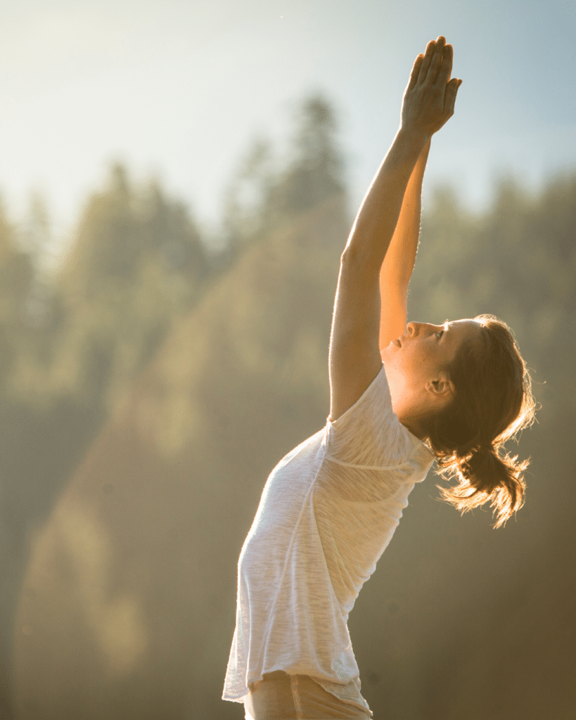 Femme les bras joints levés vers le ciel, visage renversé, lumière dorée en forêt – BYOU L'Expérience
